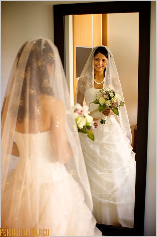 Noiva pronta de véu na cabeça  e bouquet na mão, num espelho.