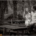Sentada nas costas do banco de uma mesa na floresta da Peninha, a noiva com o noivo junto dela, olhando um para o outro sorrindo, com o fotógrafo de casamento.