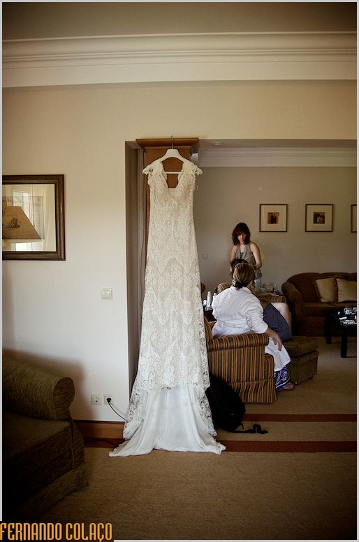 Vestido da noiva pendurado numa coluna da sala, com pessoal ao fundo.