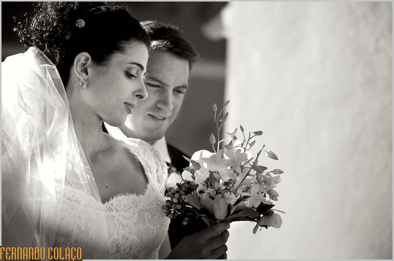 O casal olha para o bouquet da noiva.