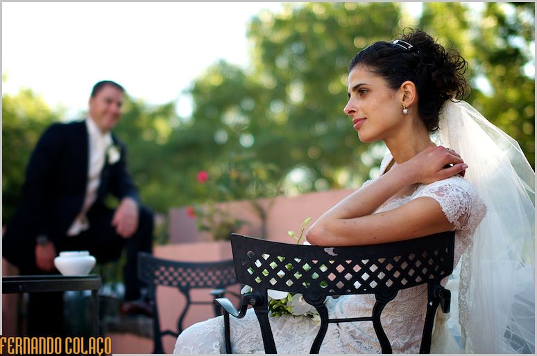 Noiva sentada numa cadeira de jardim, com o noivo, desfocado, a olhar par ela.