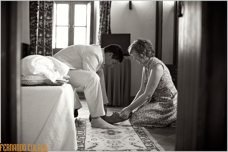 Noivo sentado e a sua mãe, sentada no chão, que o ajuda a calçar os sapatos.