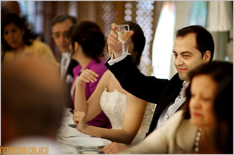 The groom, seated at the wedding meal table, makes a toast with a glass in his hand.