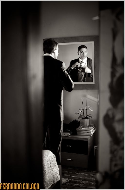 Noivo, de pé, em frente a um espelho na parede, por cima de um móvel.