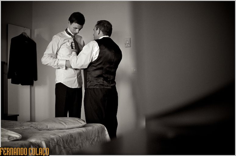 Sob a luz que vem da janela, o noivo é  ajudado pelo pai coma gravata.
