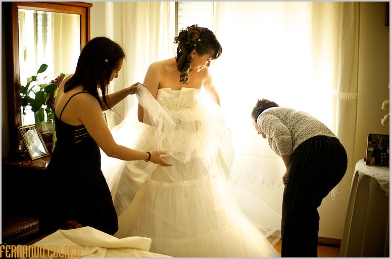 A mãe de noiva verifica se o vestido está bem, com o auxílio da irmã, enquanto ela olha para baixo.