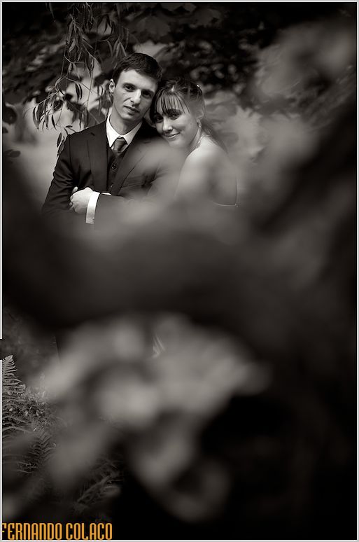 Between the trunks of a tree in the garden, the bride and groom look at the wedding photographer, in a black and white portrait.