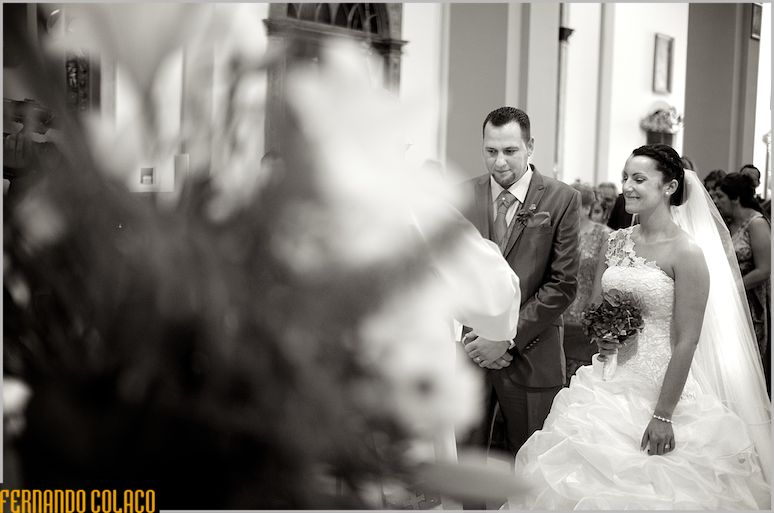 Listening to the priest, among blurred flowers, the couple smiles after the wedding ceremony.