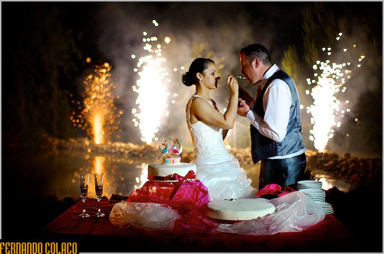 With fireworks behind, the bride and groom taste the wedding cake.