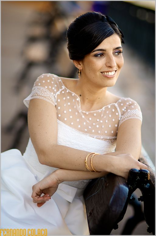 The bride sitting on a garden bench and leaning over it, looks to the side.