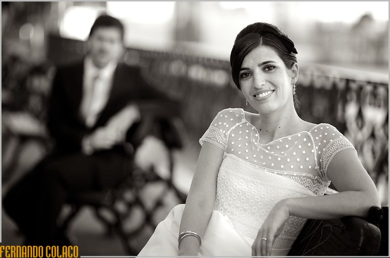 The bride, sitting on a garden bench with the groom, out of focus, in the background sitting on another.