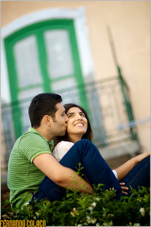 Entre as flores do muro e uma janela desfocada, o noivo beija a noiva na face.