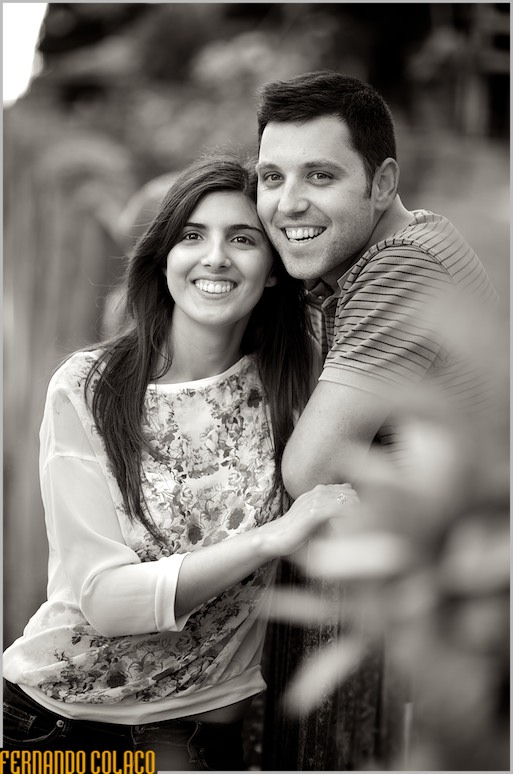 Junto a muro de ferro forjado, os noivos, juntos, olham para longe, na sessão de namoro com o fotógrafo de casamento.