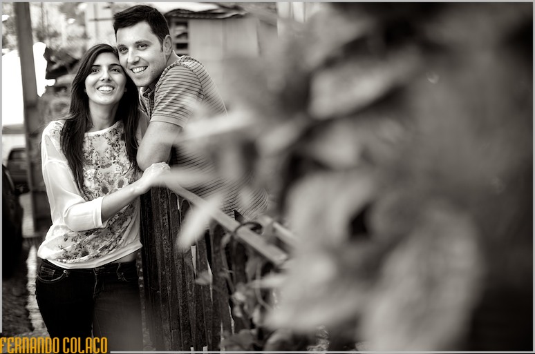 Com um tufo de flores desfocadas em primeiro plano, o casal, feliz, junto de um muro de ferro.