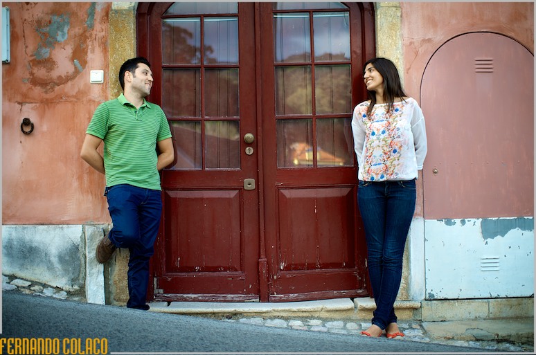 Junto de uma parede com uma porta antiga, o casal, na sessão de pré-casamento, olha um para o outro.