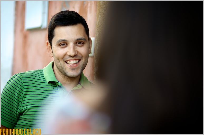 O noivo, sorrindo, olha para a noiva à sua frente, desfocada, na sessão de pré-casamento.