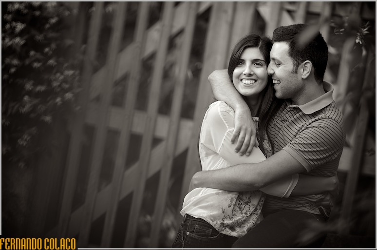 O noivo abraça noiva, rindo para ela, em frente de um portão de madeira, na sessão de pré-casamento.