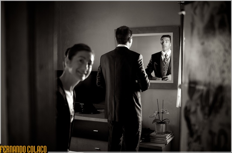 Num espelho na parede, o noivo verifica se está tudo bem no fato, já vestido, junto da irmã.