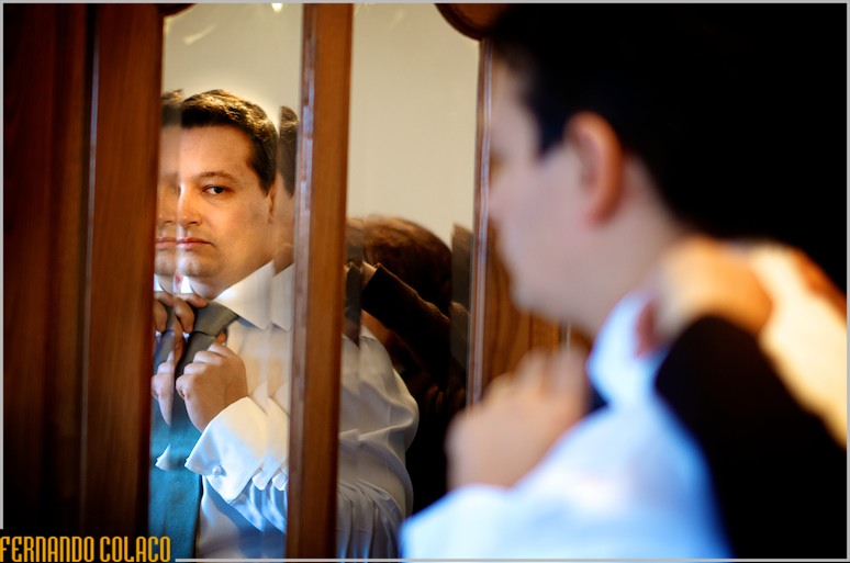 Noivo, num espelho com efeitos visuais, aperta o nó da gravata, com a mãe ajudando por detrás.