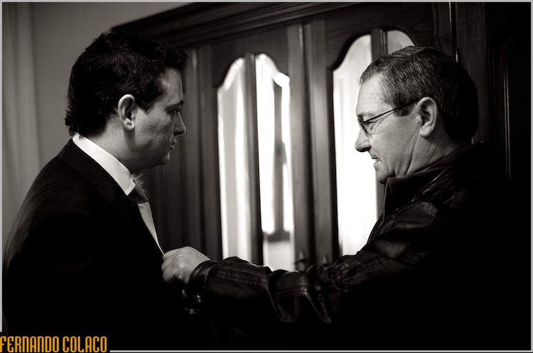 Em frente a um móvel com espelho, o pai do noivo abotoa-lhe o casaco.
