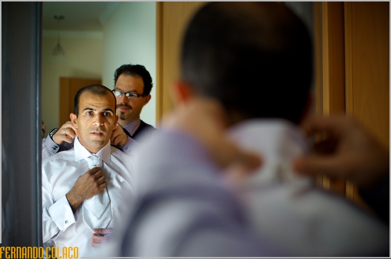 The groom, with a friend helping him with his tie, in a mirror.
