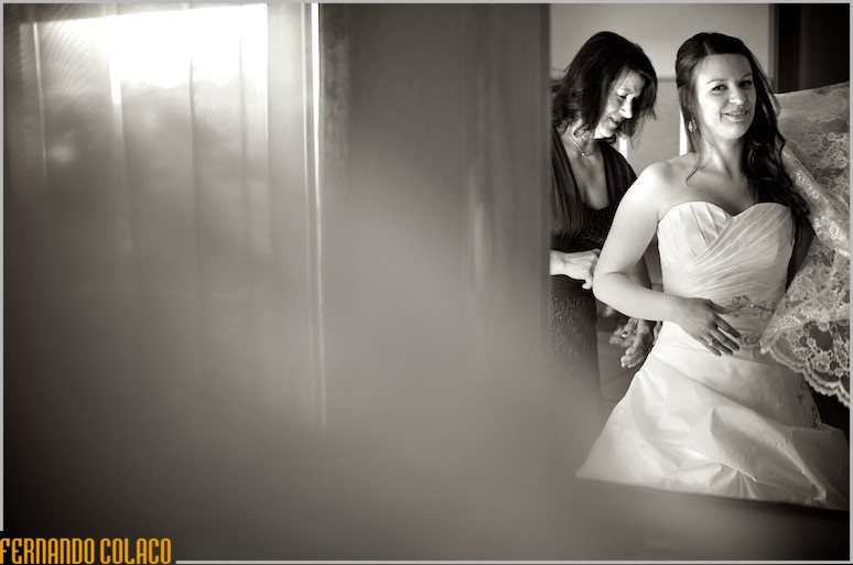 Through a window, the bride with her mother tightening her dress.