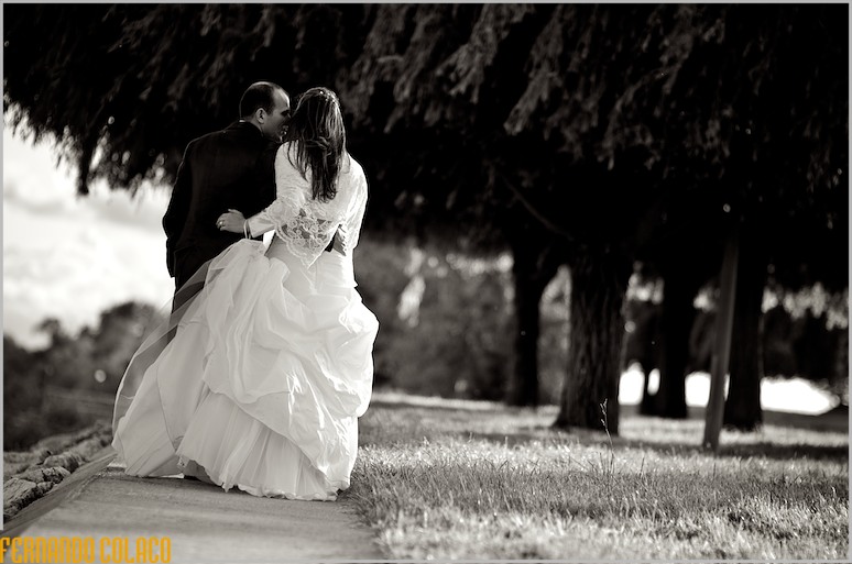 On a garden path, with large trees, the bride and groom walk together, seen from behind.
