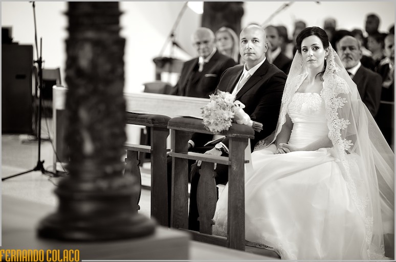 Os noivos sentados, olhando para o altar, onde estão os padres que oficiam a cerimónia do casamento.