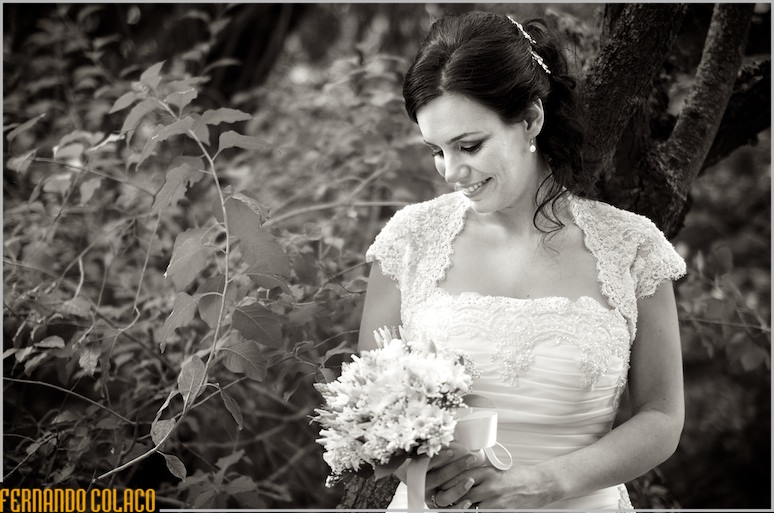 Junto de uma árvore e arbustos, a noiva olha para o bouquet que tem nas mãos.
