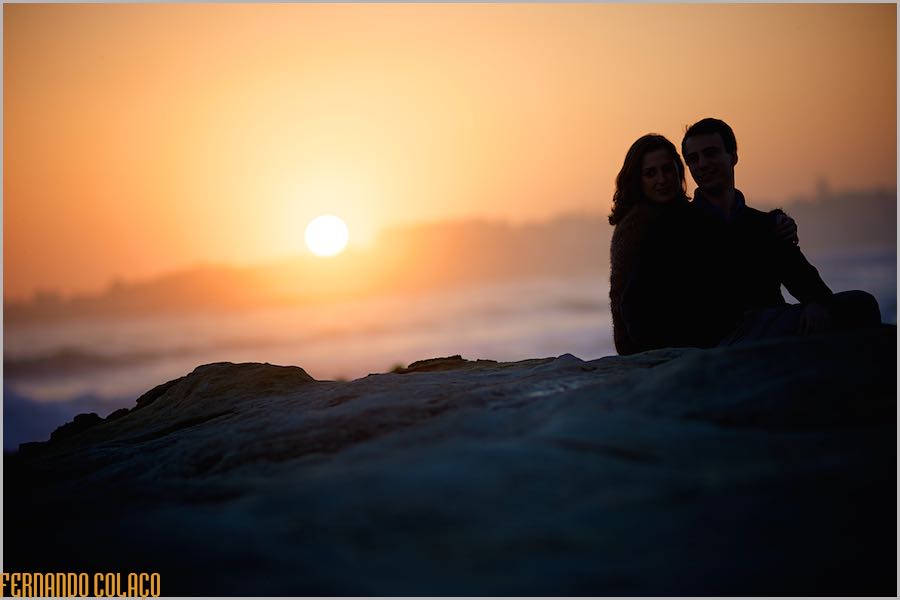 Silhueta dos noivos sobre um rochedo na praia do Estoril, com a cor laranja do sol, quando se está a pôr sobre Cascais.