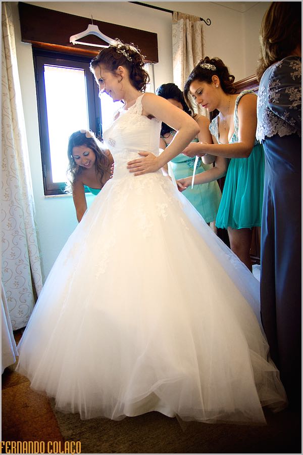A noiva, de pé, em frente de amigas que a ajudam no vestir do vestido.