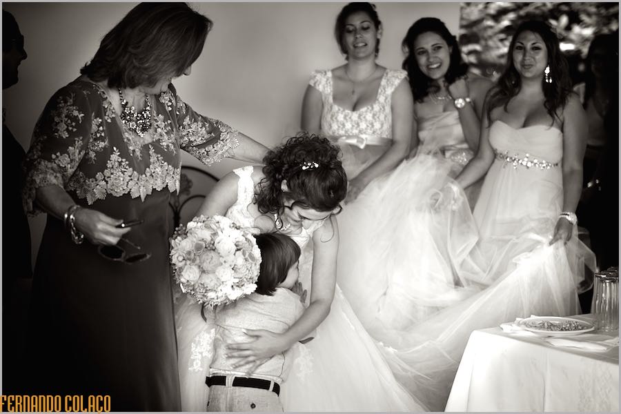 Em frente de amigas que lhe seguram na cauda do vestido, a noiva, com o bouquet na mão, abraça um menino.