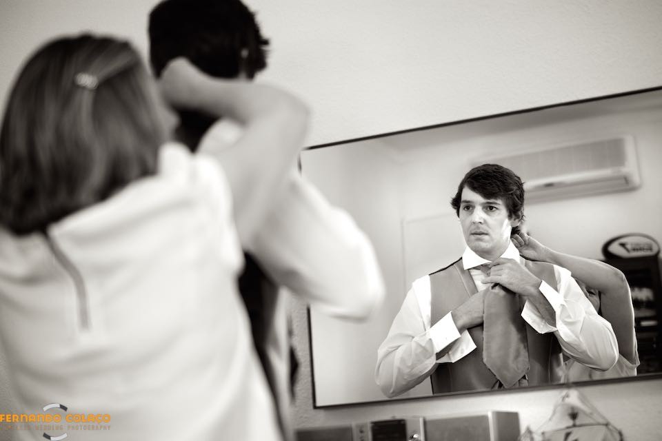 Num espelho, em frente deles, o noivo aperta o nó da gravata com a ajuda da mãe.