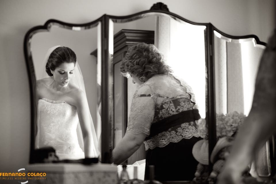 Em casa dos pais, a noiva aparece num espelho com a mãe que lhe faz os últimos acertos no vestido.