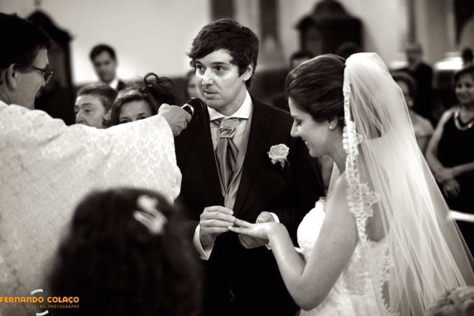 O noivo no juramento do casamento enquanto põe a aliança no dedo da noiva, a seu lado.