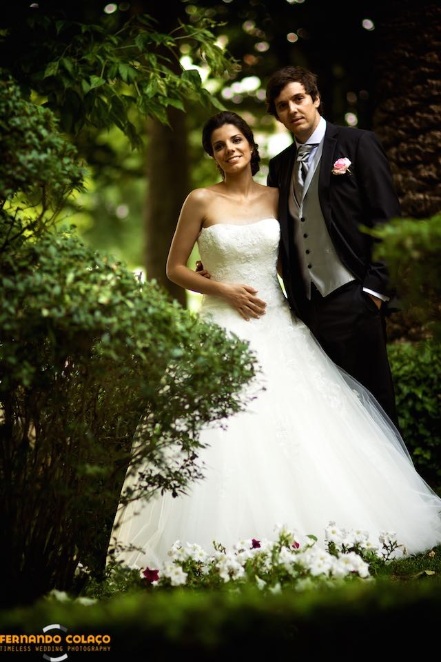 De pé e juntos, os noivos em pose clássica, entre as plantas do jardim, para o fotógrafo de casamento.