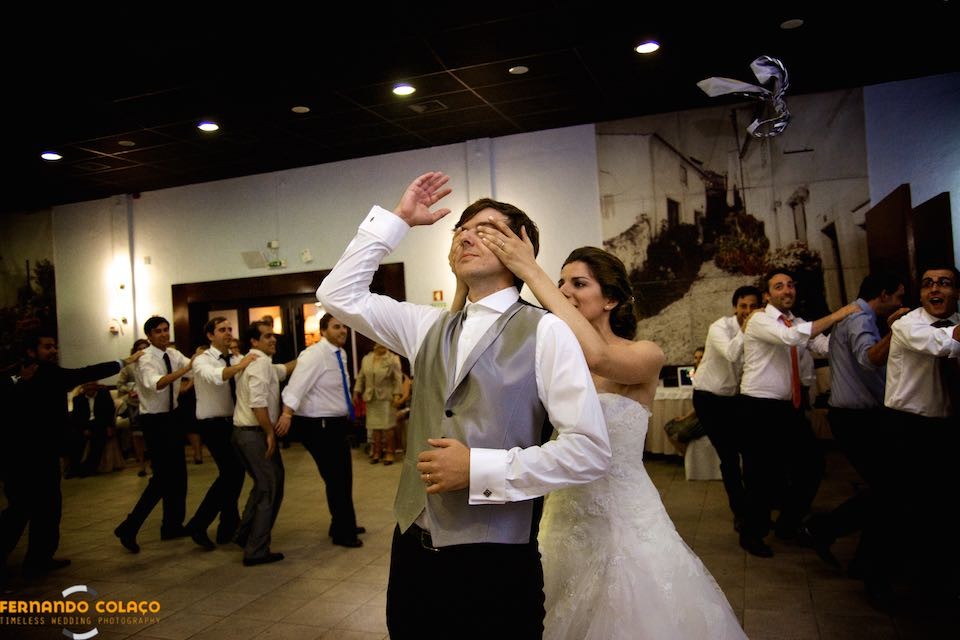 Num jogo da festa do casamento, a noiva tapa com as mãos os olhos do noivo com convidados dançando por detrás.