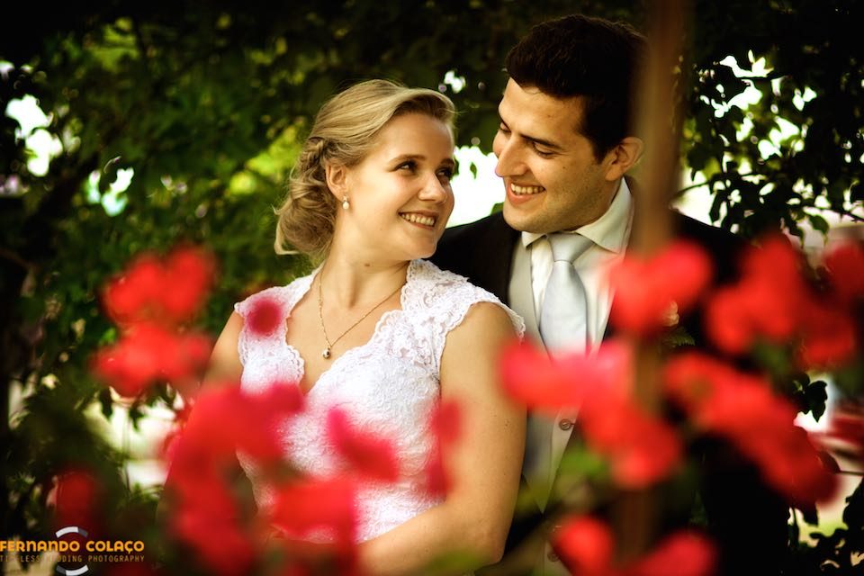 Among red flowers, the bride and groom smile together.