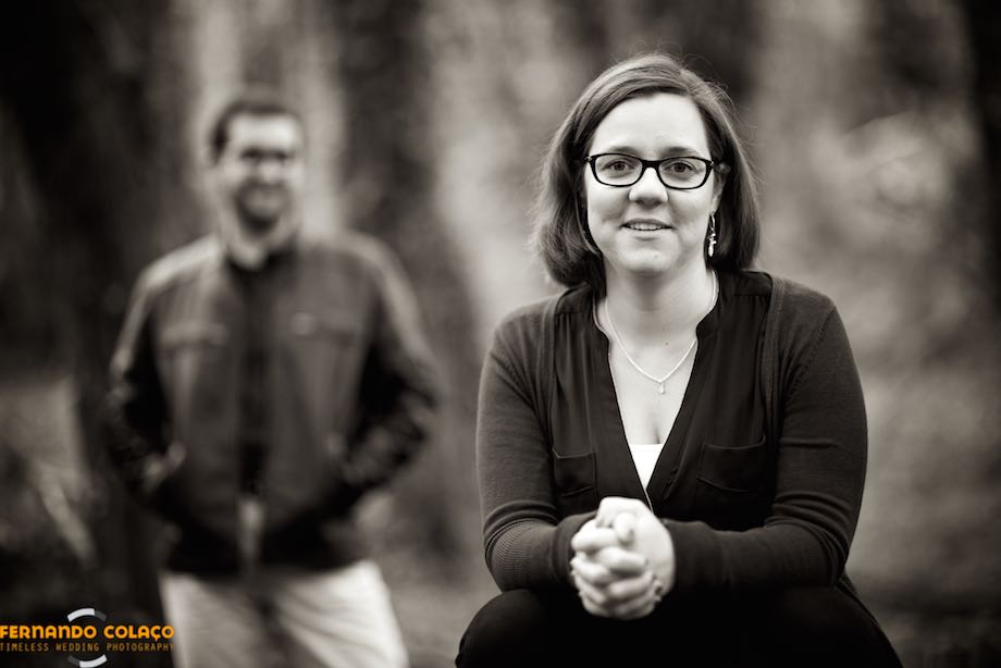 The bride, seated and with the groom behind, out of focus, looks at the wedding photographer, in the courtship session.
