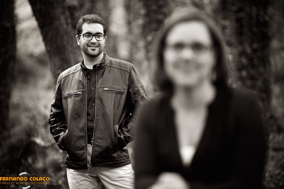 Looking at the bride, seated in front of him and out of focus, the groom, hands in his coat pockets, smiles during the pre-wedding session.