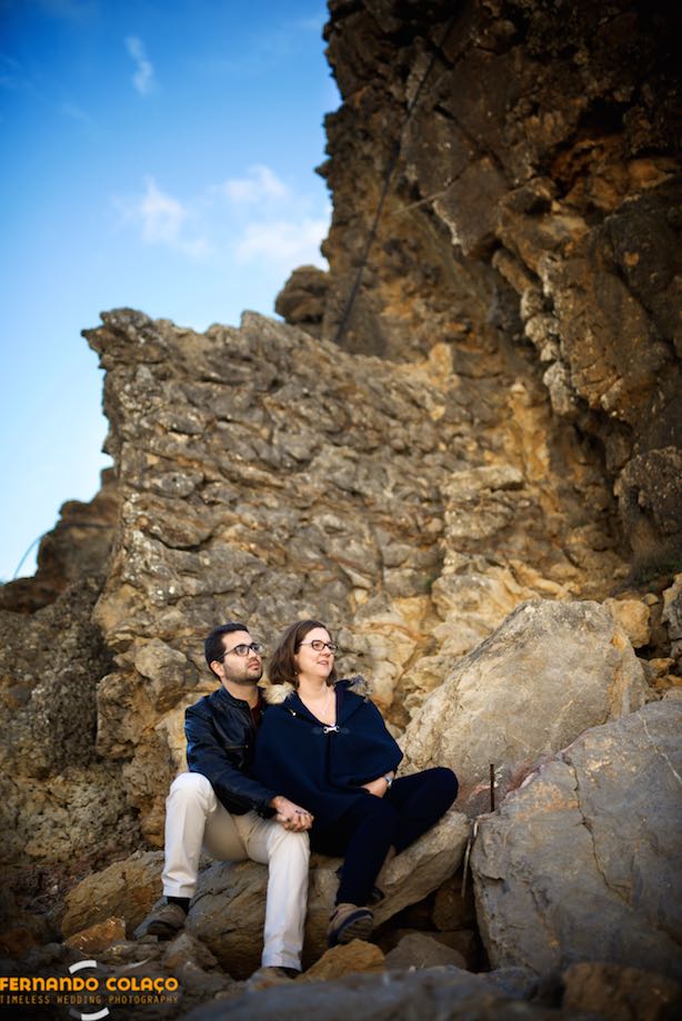 Sitting on a rock at Guincho beach in Cascais, the bride and groom pose for the wedding photographer by the cliffs.