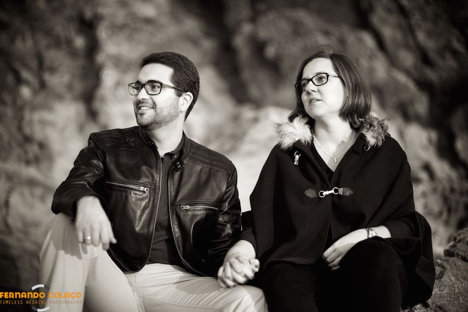The couple in a pre-wedding session, sitting on a cliff at Guincho beach, seen from the front by the wedding photographer.