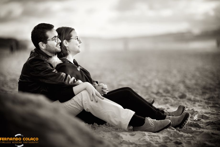 In the late afternoon light on Guincho beach in Cascais, the bride leans against the groom's chest, sitting on the sand.