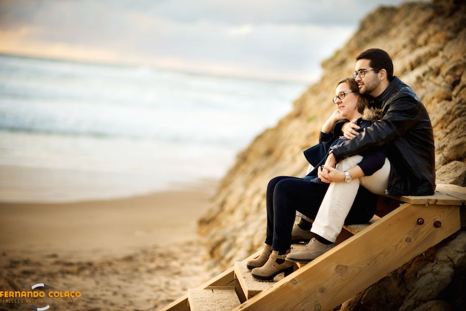 On a wooden staircase next to the beach, the bride and groom enjoy the sunset, seated, during the courtship session with the wedding photographer.