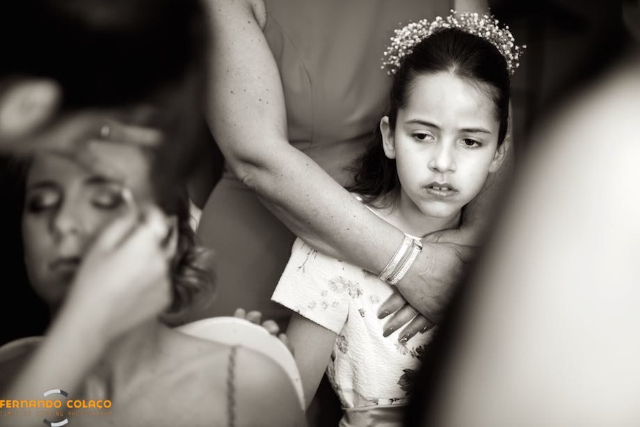 Enquanto a noiva é maquilhada, em primeiro plano e desfocada, uma menina assiste muito atenta.