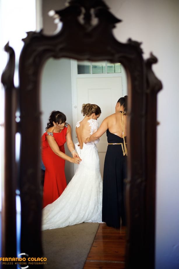 Num espelho grande de sala, a noiva, de costas, no meio de duas amigas que lhe abotoam o vestido.