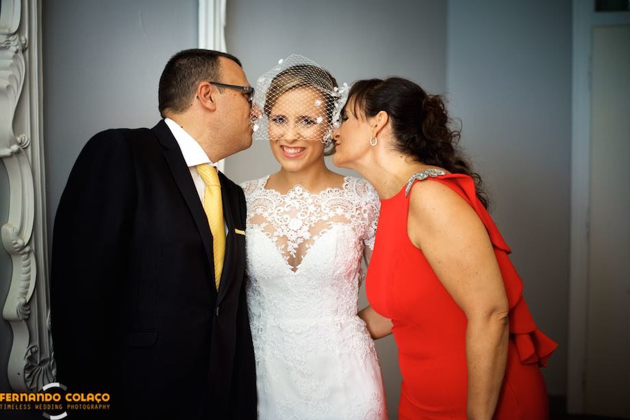 Já pronta para ir para a cerimónia do casamento, os pais da noiva beijam-na na face ao mesmo tempo, um de cada lado.