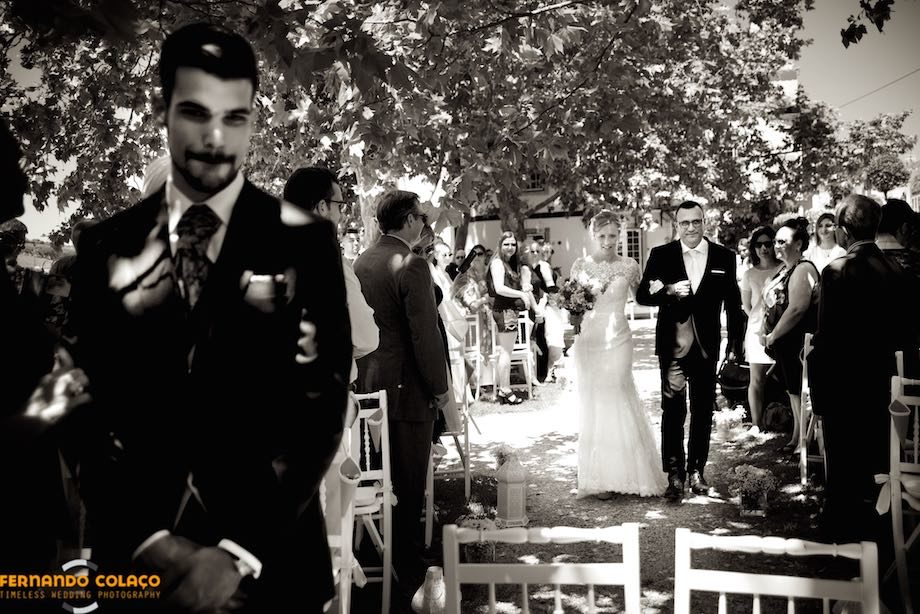 Com o noivo em primeiro plano, a noiva percorre o caminho, entre convidados, na direcção da mesa cerimonial.