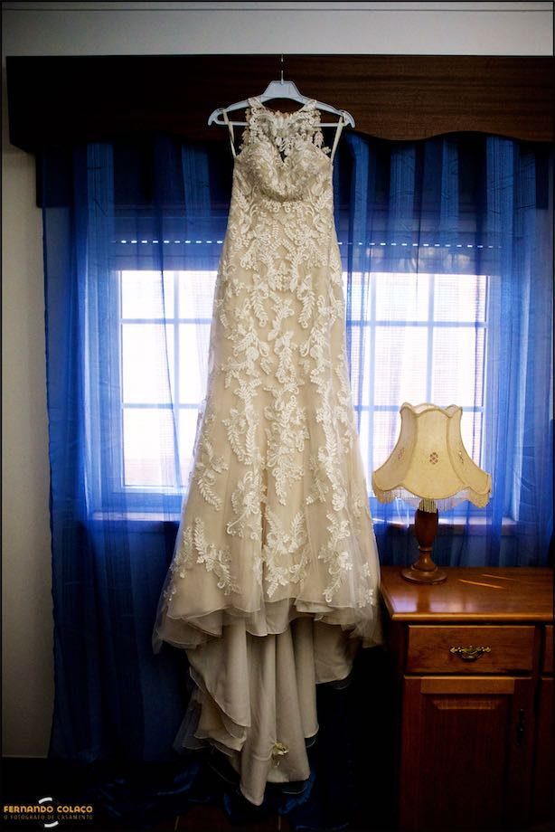 Wedding dress in front of blue curtained window and a lamp on a table.