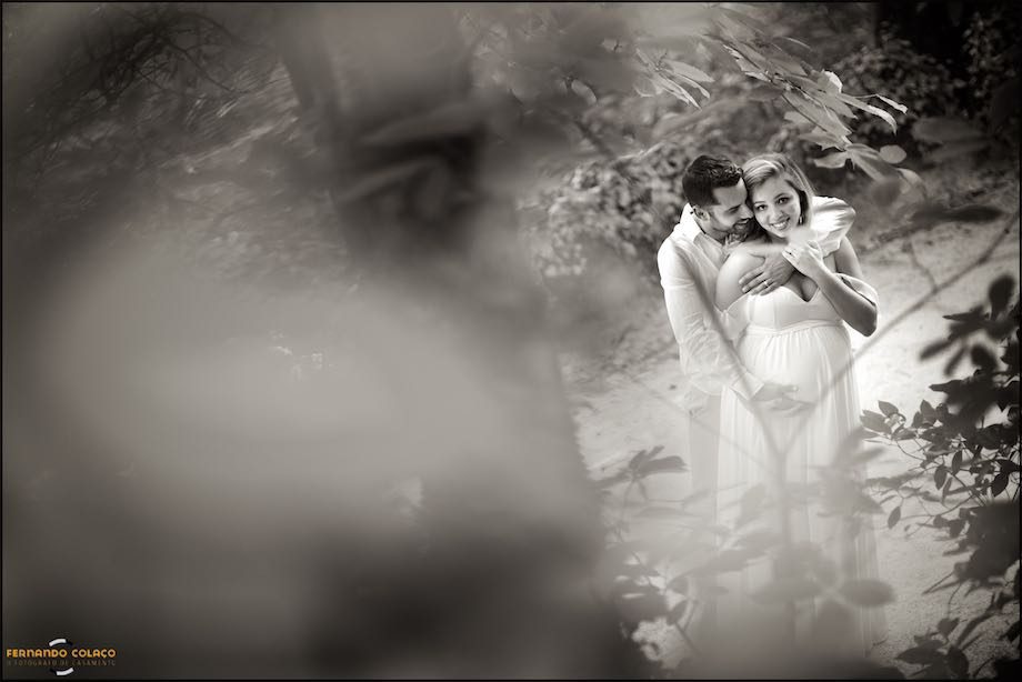 Marido abraça a sua mulher grávida, por detrás, entre árvores folhosas.